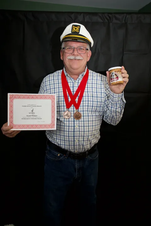 Charlie Adams in a captain's hat wearing two 2022 Scovie Awards medals, holding the Scovie certificate for 3rd Place, Barbecue Sauce, Mustard-Hot and a jar of Grandpa Spencer's Hot & Spicy Mustard