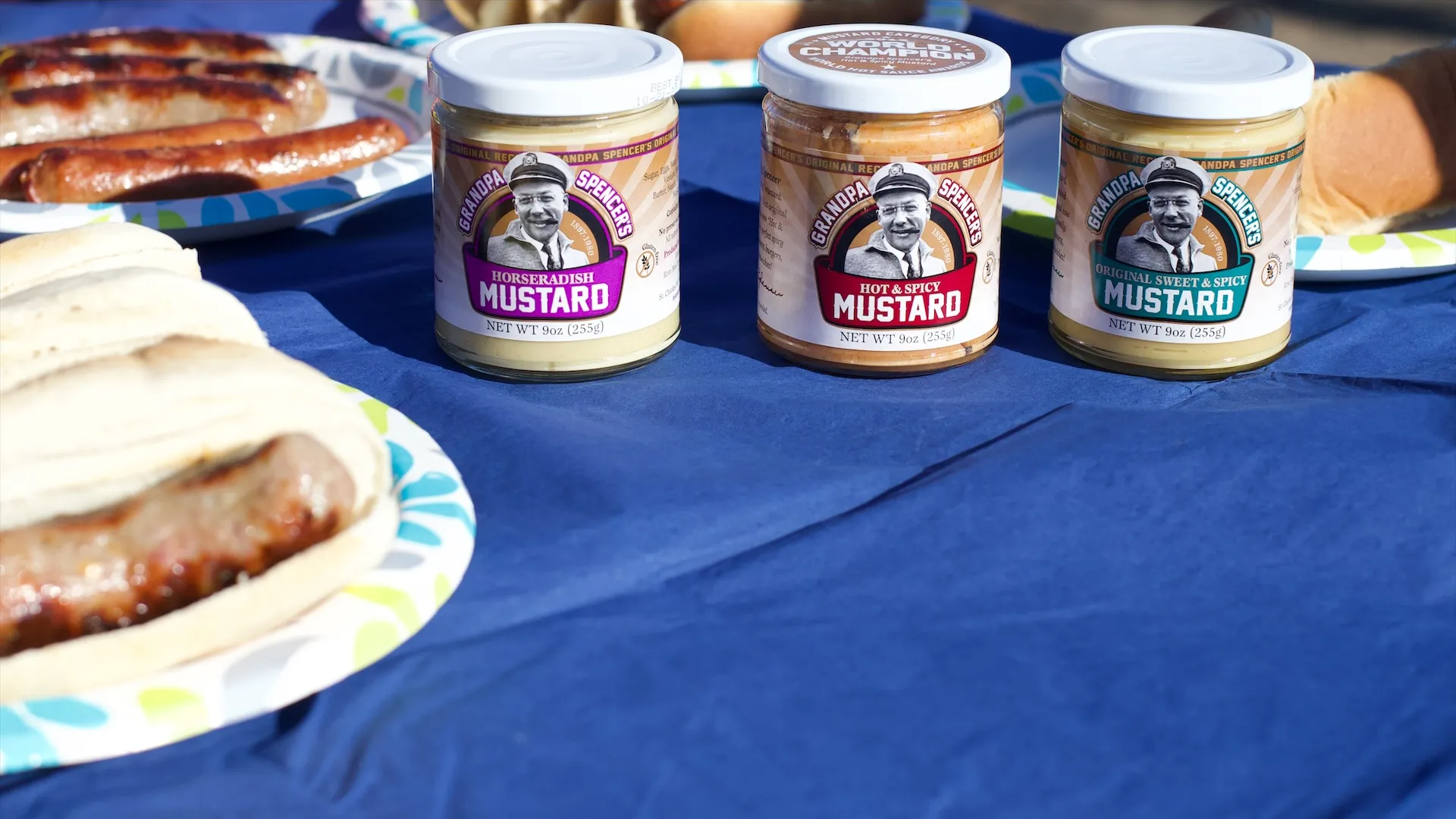 Grandpa Spencer's Horseradish, Hot & Spicy and Original Sweet & Spicy mustard jars on a blue picnic table next to plates of grilled brats in buns; the Hot & Spicy lid shows its World Champion seal