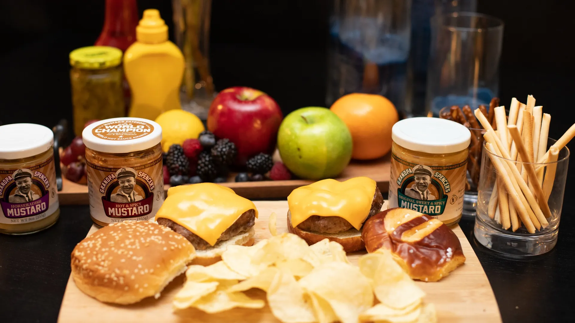 Two cheeseburgers, a sesame bun, a pretzel bun and potato chips on a wooden board, with jars of Grandpa Spencer's Horseradish, Hot & Spicy and Original Sweet & Spicy mustard behind them; the Hot & Spicy lid shows its World Champion seal
