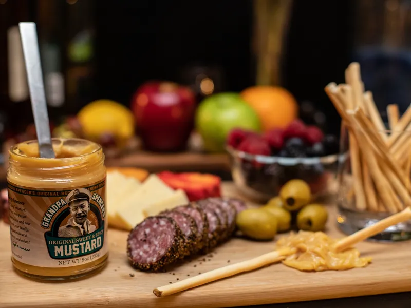 An open jar of Grandpa Spencer's Original Sweet & Spicy Mustard with a spoon in it, on a wooden board with sliced salami, white cheese, green olives and breadsticks